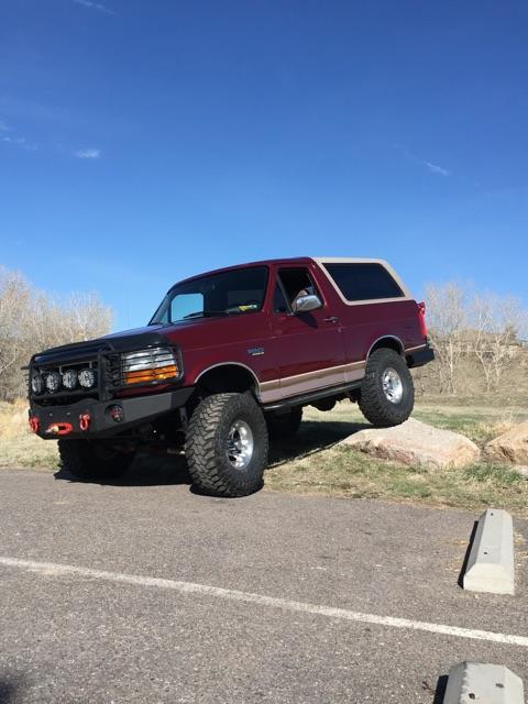 1996 Ford Bronco - A maroon 1996 Ford Bronco with a tan top is parked on a slight incline with its front tires on a rock. The vehicle has a custom front bumpe…