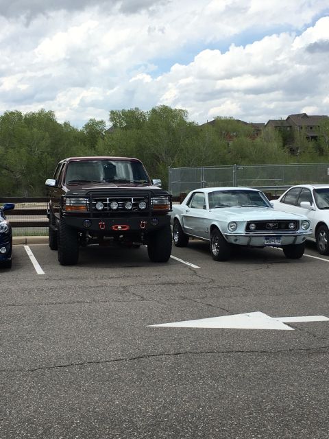 1996 Ford Bronco - A lifted, dark red 1996 Ford Bronco is parked next to a white classic Ford Mustang in a parking lot with a white directional arrow painted…