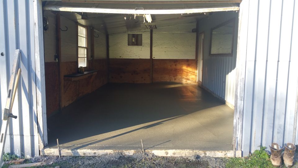 Freshly poured concrete floor inside a shed with tools and boots outside.