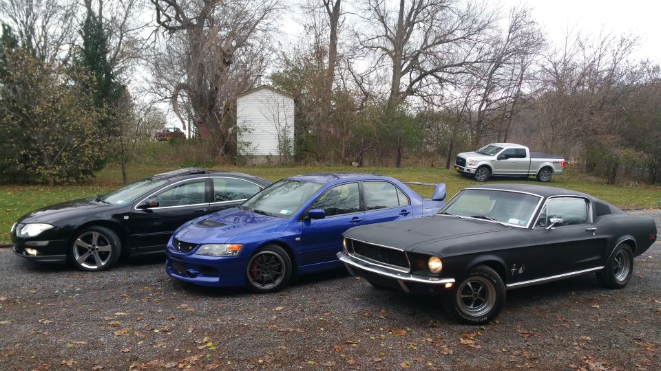 Black 1968 Ford Mustang fastback parked on gravel, with other cars and a pickup truck in the background.