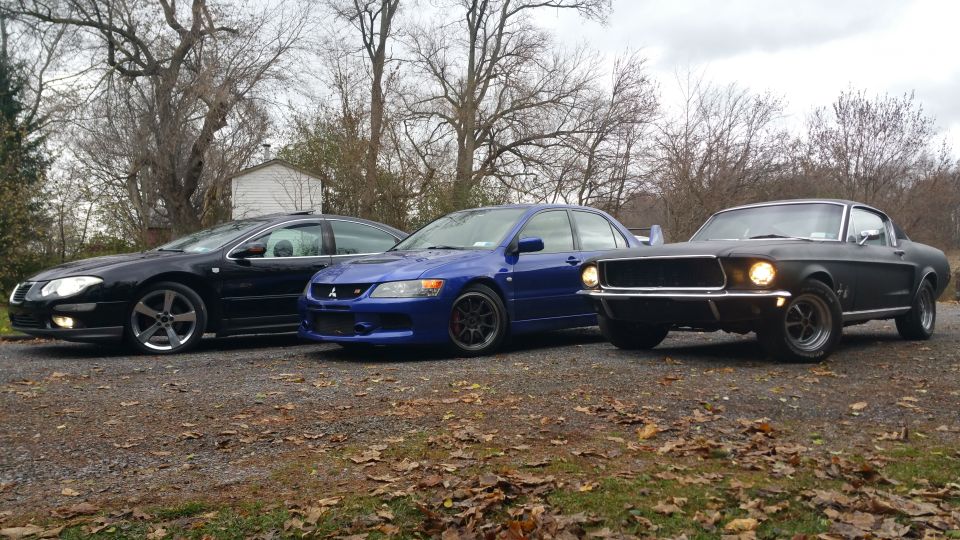 Three cars parked on a gravel driveway: a black sedan, a blue performance sedan, and a dark gray classic coupe.