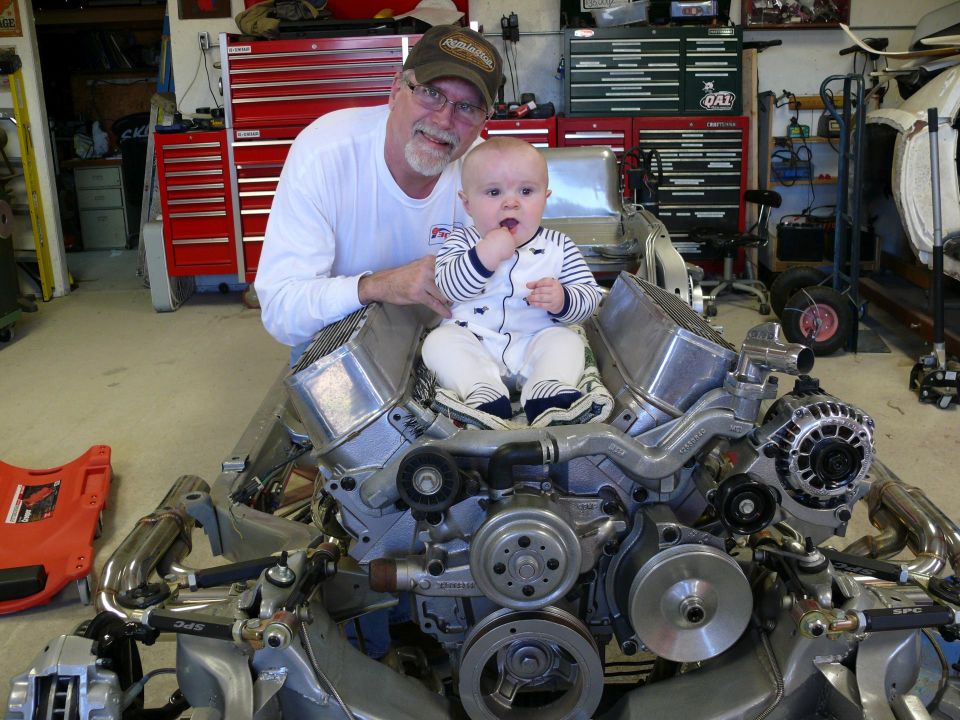 Man and baby on top of a V8 engine with polished aluminum parts in a garage.