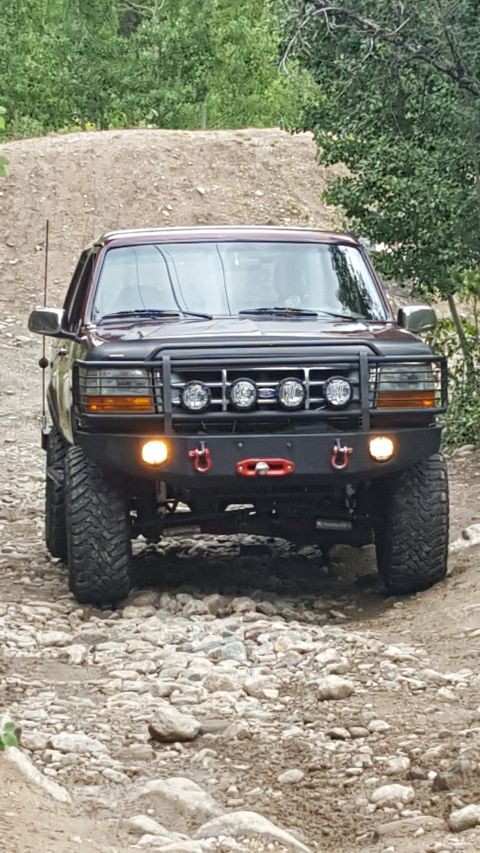 1996 Ford Bronco - A dark red 1996 Ford Bronco is shown from the front, driving on a rocky, off-road trail. The vehicle features a custom bumper with a winch…