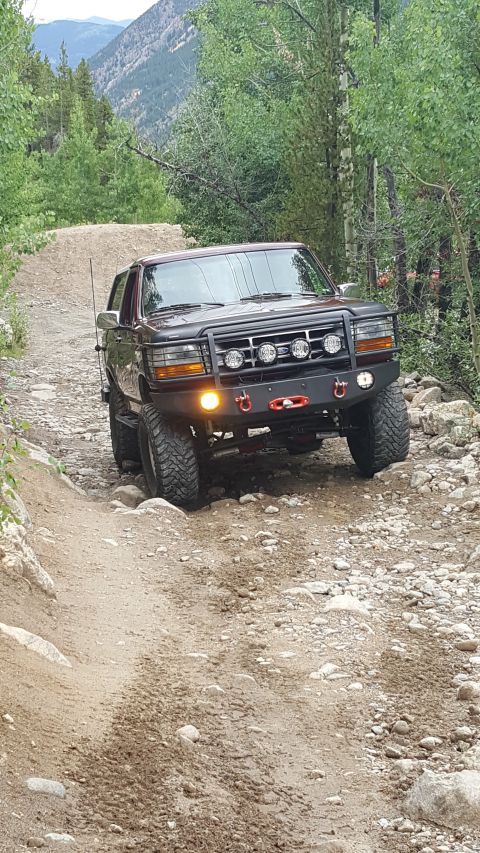 1996 Ford Bronco - A dark red 1996 Ford Bronco is navigating a rocky, off-road trail surrounded by trees and mountains. The Bronco features a custom front bum…