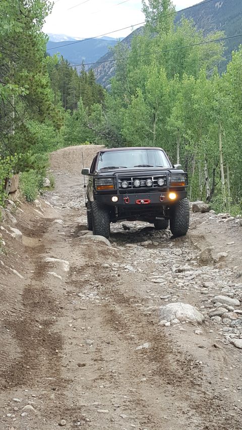 1996 Ford Bronco - A black Ford Bronco is driving on a rocky, dirt trail through a forest with mountains in the background. The vehicle has a custom front bum…