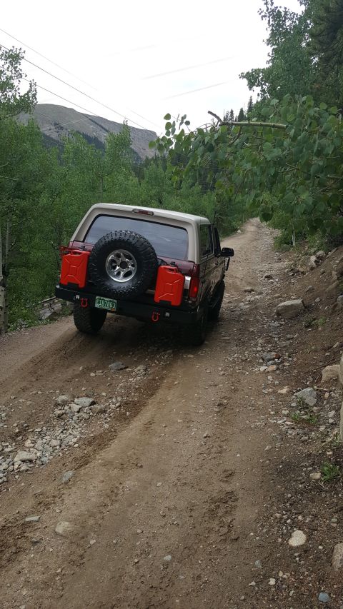 1996 Ford Bronco - The rear view of a maroon and tan 1996 Ford Bronco is shown driving on a rocky, dirt road surrounded by trees. The vehicle has a spare tire…