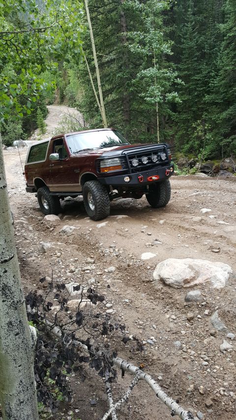 1996 Ford Bronco - A maroon 1996 Ford Bronco is shown from a 3/4 front angle on a dirt road in a forest. The vehicle has a custom front bumper with multiple l…