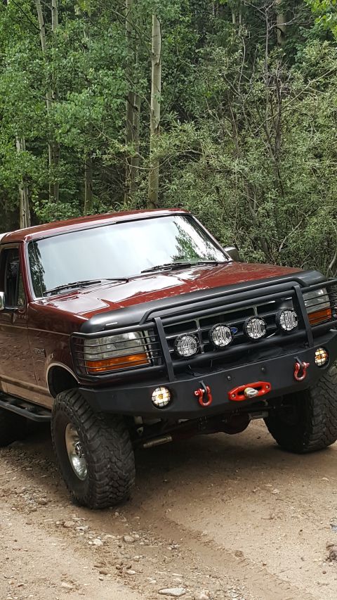 1996 Ford Bronco - A maroon 1990s Ford Bronco is parked on a dirt road with trees in the background. The front of the SUV features a black bull bar with four…