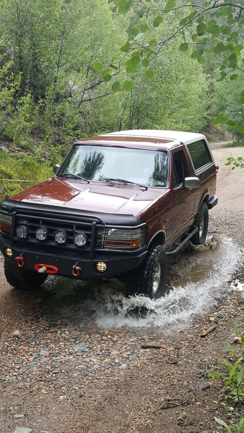 1996 Ford Bronco - A maroon 1996 Ford Bronco is driving through shallow water on a dirt road surrounded by trees. The vehicle has a black brush guard with KC…