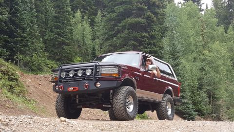 1996 Ford Bronco - A maroon 1996 Ford Bronco is parked on a gravel road in a forest, with a person visible in the driver's seat giving a thumbs up. The Bronco…
