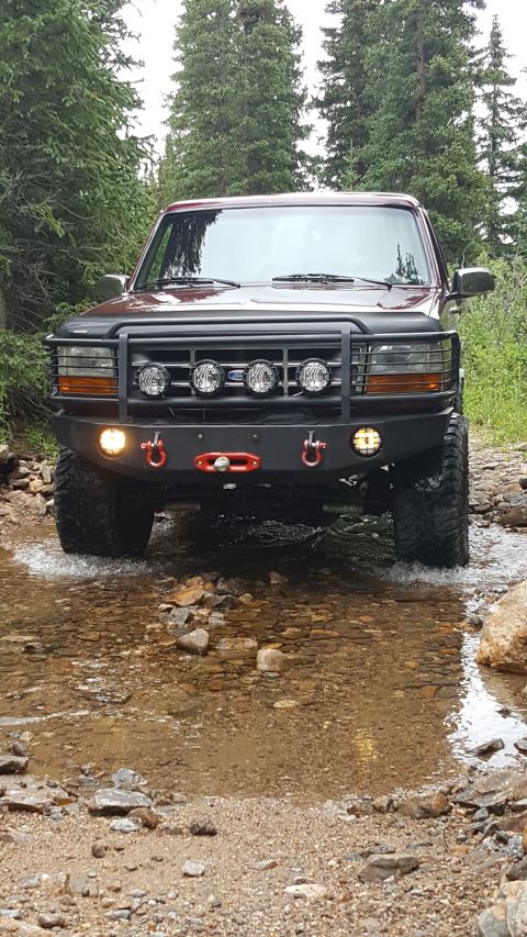 1996 Ford Bronco - A dark red 1996 Ford Bronco is shown from the front, driving through a shallow, rocky stream in a forest.