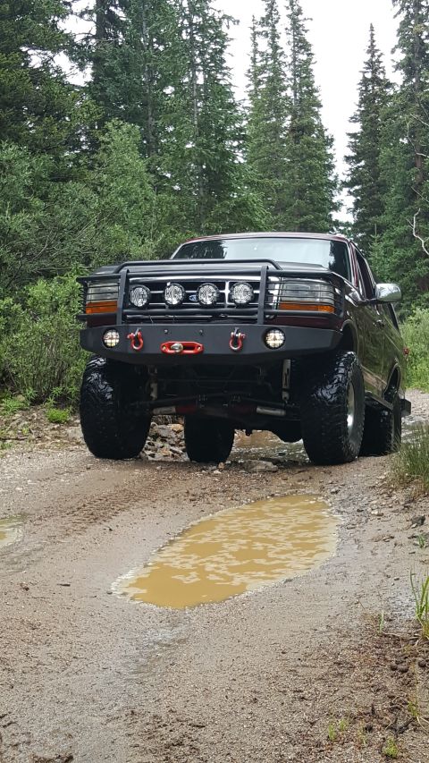 1996 Ford Bronco - A lifted, dark red 1996 Ford Bronco with an aftermarket bumper and KC lights drives on a muddy, wooded trail with puddles.