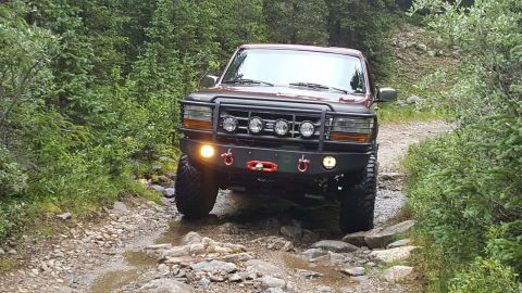 1996 Ford Bronco - A dark red Ford Bronco is driving on a rocky, wet trail surrounded by lush green trees. The vehicle has an aftermarket bumper with multiple…