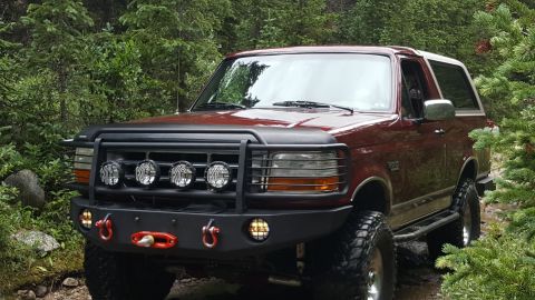 1996 Ford Bronco - A maroon 1996 Ford Bronco is parked on a dirt path surrounded by dense green trees. The front of the vehicle features a black bull bar with…