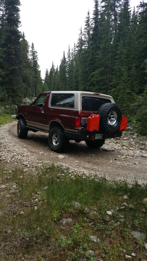 1996 Ford Bronco - A maroon and tan 1996 Ford Bronco is parked on a rocky, dirt road surrounded by tall pine trees. The rear of the vehicle shows a spare tire…