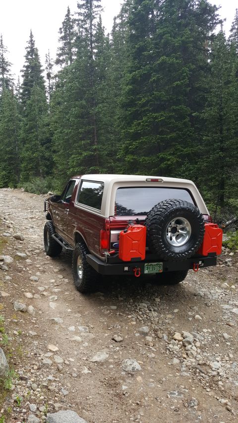 1996 Ford Bronco - A maroon and tan 1996 Ford Bronco is parked on a rocky dirt road surrounded by a dense forest of evergreen trees. The rear of the SUV featu…