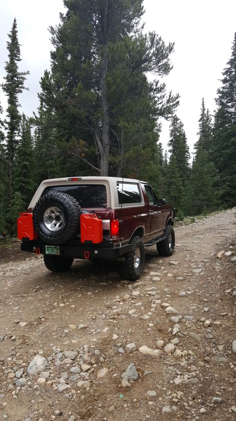 1996 Ford Bronco - The rear driver's side view of a maroon 1996 Ford Bronco is parked on a rocky dirt road surrounded by pine trees. The vehicle has a spare t…