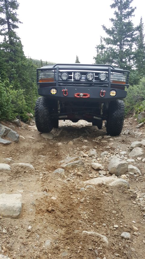 1996 Ford Bronco - The front view of a modified dark-colored SUV is shown navigating a rocky, dirt trail in a wooded area. The vehicle features a heavy-duty b…