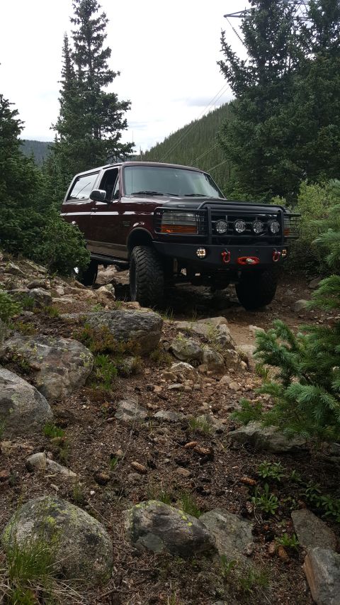 1996 Ford Bronco - A maroon Ford Bronco is navigating a rocky, wooded trail. The SUV is equipped with an aftermarket front bumper, winch, and auxiliary lights.