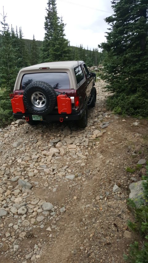 1996 Ford Bronco - A maroon 1996 Ford Bronco with a tan top is parked on a rocky, wooded trail. The rear of the SUV features a spare tire and two red jerry ca…