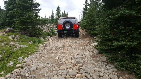 1996 Ford Bronco - The rear of a tan SUV is shown driving up a rocky, mountainous trail surrounded by pine trees. Two red jerry cans are mounted on the rear b…