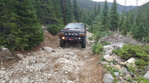 1996 Ford Bronco - A dark red 1996 Ford Bronco is driving on a rocky, off-road trail surrounded by pine trees. The Bronco features an aftermarket front bumper…