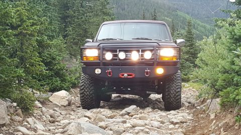 1996 Ford Bronco - A dark-colored Ford Bronco is shown from the front on a rocky mountain trail. The vehicle has a custom front bumper with multiple lights an…