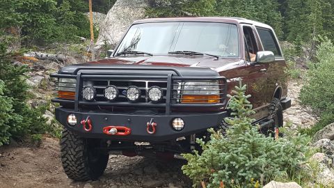 1996 Ford Bronco - A dark red 1996 Ford Bronco is parked on a rocky, wooded trail. The front of the vehicle features a black brush guard with four KC lights a…