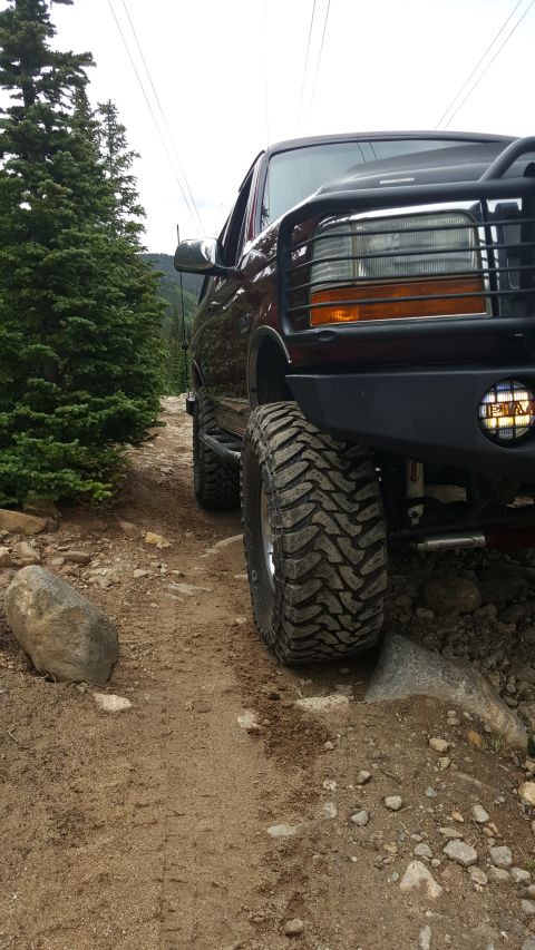 1996 Ford Bronco - A dark-colored Ford Bronco is shown from a low-angle front-three-quarter view, driving on a rocky, dirt trail surrounded by trees. The vehi…