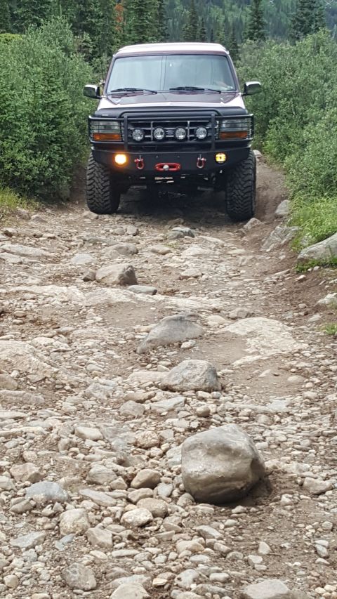 1996 Ford Bronco - A maroon 1996 Ford Bronco is driving up a rocky, off-road trail surrounded by dense green foliage. The front of the SUV features a heavy-du…