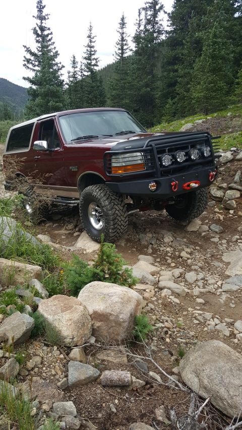 1996 Ford Bronco - A maroon 1996 Ford Bronco with a white top is parked on a rocky, off-road trail surrounded by pine trees.