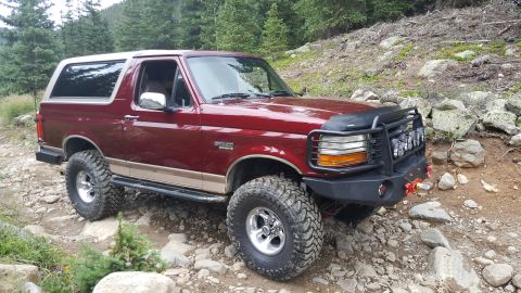 1996 Ford Bronco - A red 1996 Ford Bronco with a tan stripe is parked on a rocky, wooded trail. The SUV features a custom black front bumper with integrated l…