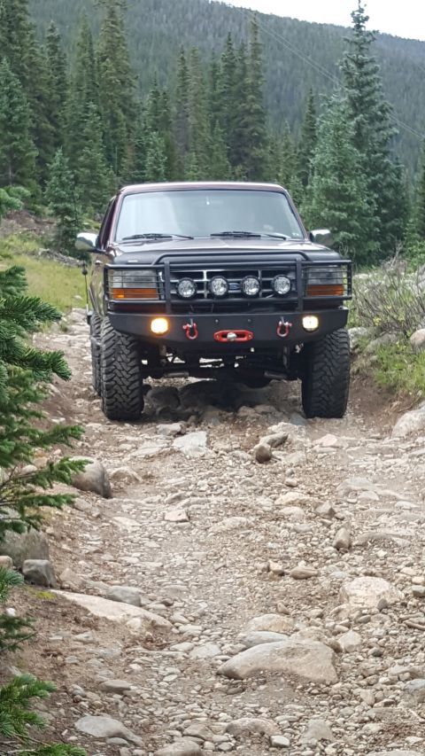 1996 Ford Bronco - A dark colored Ford Bronco is driving on a rocky, off-road trail surrounded by pine trees and a mountain in the background. The vehicle has…