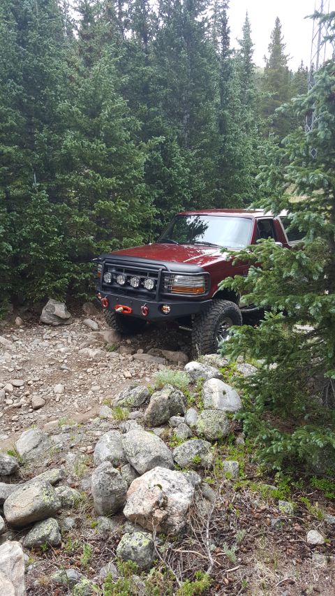 1996 Ford Bronco - A dark red Ford Bronco is parked on a rocky, wooded trail. The vehicle features an aftermarket front bumper with multiple lights and a winc…