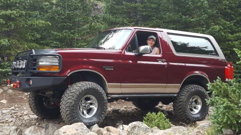 1996 Ford Bronco - A maroon 1996 Ford Bronco with a tan lower body trim is shown from the side, driving on a rocky, wooded trail. The driver, wearing a baseba…