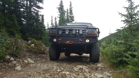 1996 Ford Bronco - A lifted 1996 Ford Bronco is shown from the front on a rocky, wooded trail. The vehicle features a custom heavy-duty bumper with integrated…