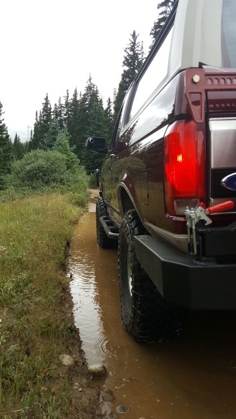 1996 Ford Bronco - The rear passenger side of a dark red Ford Bronco is shown driving through a muddy puddle on a dirt road. Tall evergreen trees line the pat…