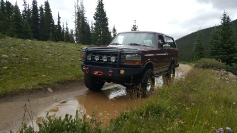 1996 Ford Bronco - A maroon 1996 Ford Bronco is driving through a muddy puddle on a dirt road in a mountainous, forested area. The Bronco is equipped with a h…