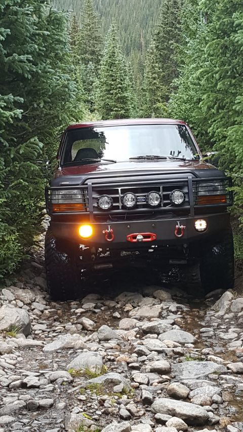 1996 Ford Bronco - A red 1996 Ford Bronco is shown from the front, navigating a rocky, wooded trail. The vehicle features an aftermarket bumper with KC lights…