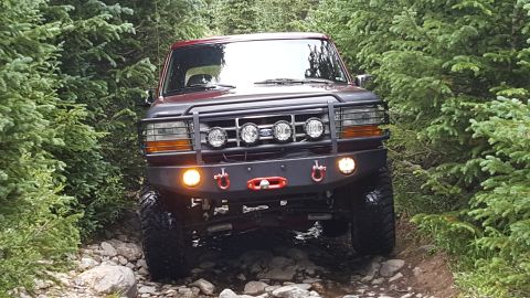 1996 Ford Bronco - A dark red 1996 Ford Bronco is driving on a rocky, wooded trail. The front of the SUV features a custom bumper with KC lights and tow hooks.