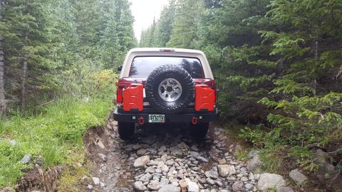 1996 Ford Bronco - The rear of a red 1996 Ford Bronco is shown driving on a rocky, wooded trail. The vehicle is equipped with a spare tire mounted on the back…