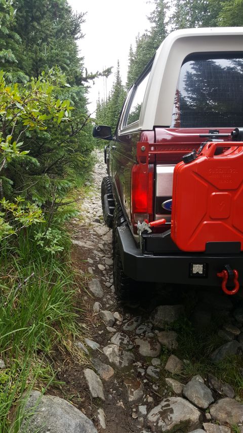 1996 Ford Bronco - The rear passenger side of a red 1996 Ford Bronco is shown on a rocky, wooded trail. A red jerry can is mounted to the rear bumper.