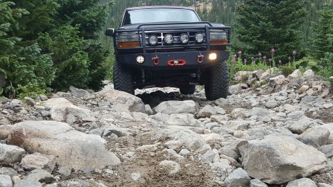 1996 Ford Bronco - A dark colored Ford Bronco is navigating a rocky, off-road trail with large boulders and pine trees visible in the background.