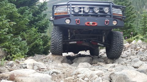 1996 Ford Bronco - The front view of a dark red Ford Bronco is shown navigating a rocky, mountainous terrain. The vehicle features a heavy-duty aftermarket bu…
