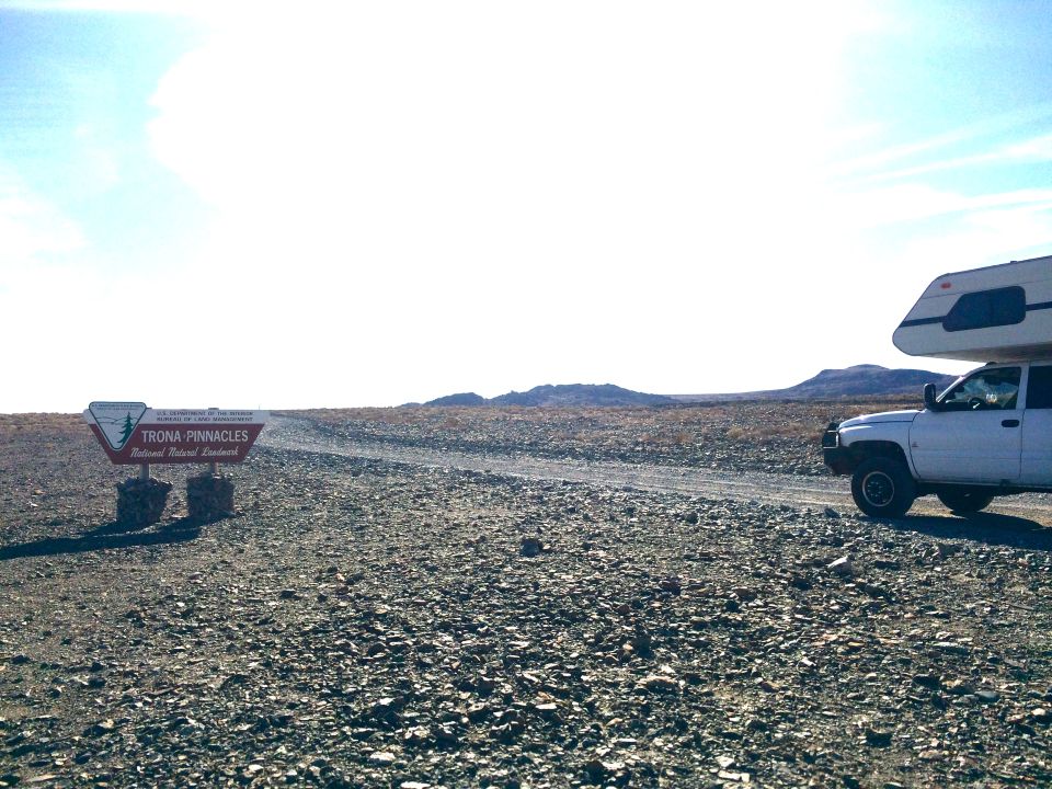 White camper truck parked on dirt road by Trona Pinnacles sign in arid landscape.