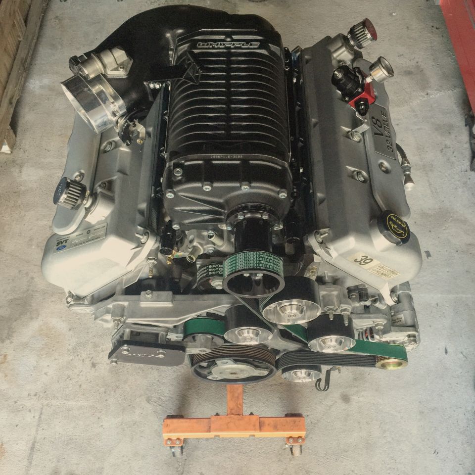 Supercharged V8 engine with black intake manifold and silver valve covers, mounted on an orange engine stand