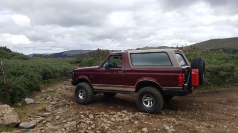 1996 Ford Bronco - 1996 Ford Bronco parked on a dirt road in a mountainous, vegetated area under a cloudy sky.
