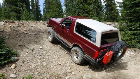 1996 Ford Bronco - A maroon Ford Bronco navigates a steep, rocky incline on a forest trail, equipped for off-road adventure.
