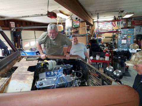 1972 Ford Bronco - Men are gathered around the engine bay of a vehicle undergoing restoration in a garage setting.
