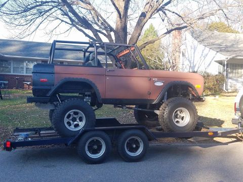 1972 Ford Bronco - A lifted 1972 Ford Bronco is transported on a trailer, its weathered brown body and roll cage visible.